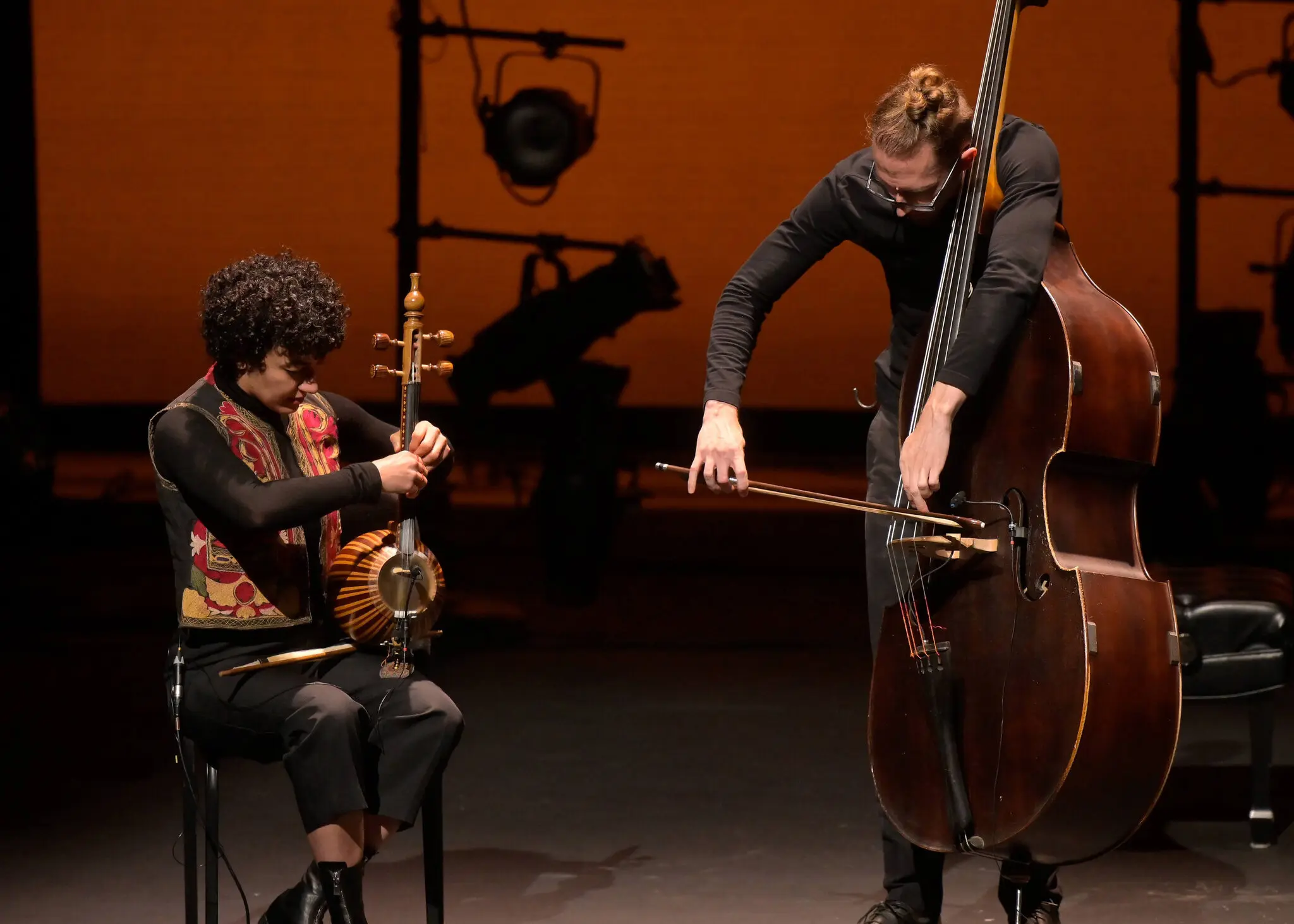 Two musicians perform on stage: one plays a bowed string instrument while seated, the other stands playing a double bass. Stage lights and a warm orange background are visible behind them.
