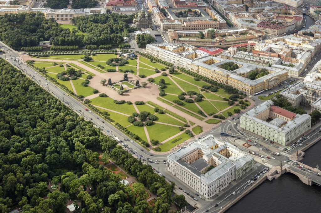Aerial view of a large, landscaped triangular park surrounded by historic buildings, tree-lined paths, and roads in a city, with a river at the bottom right corner.
