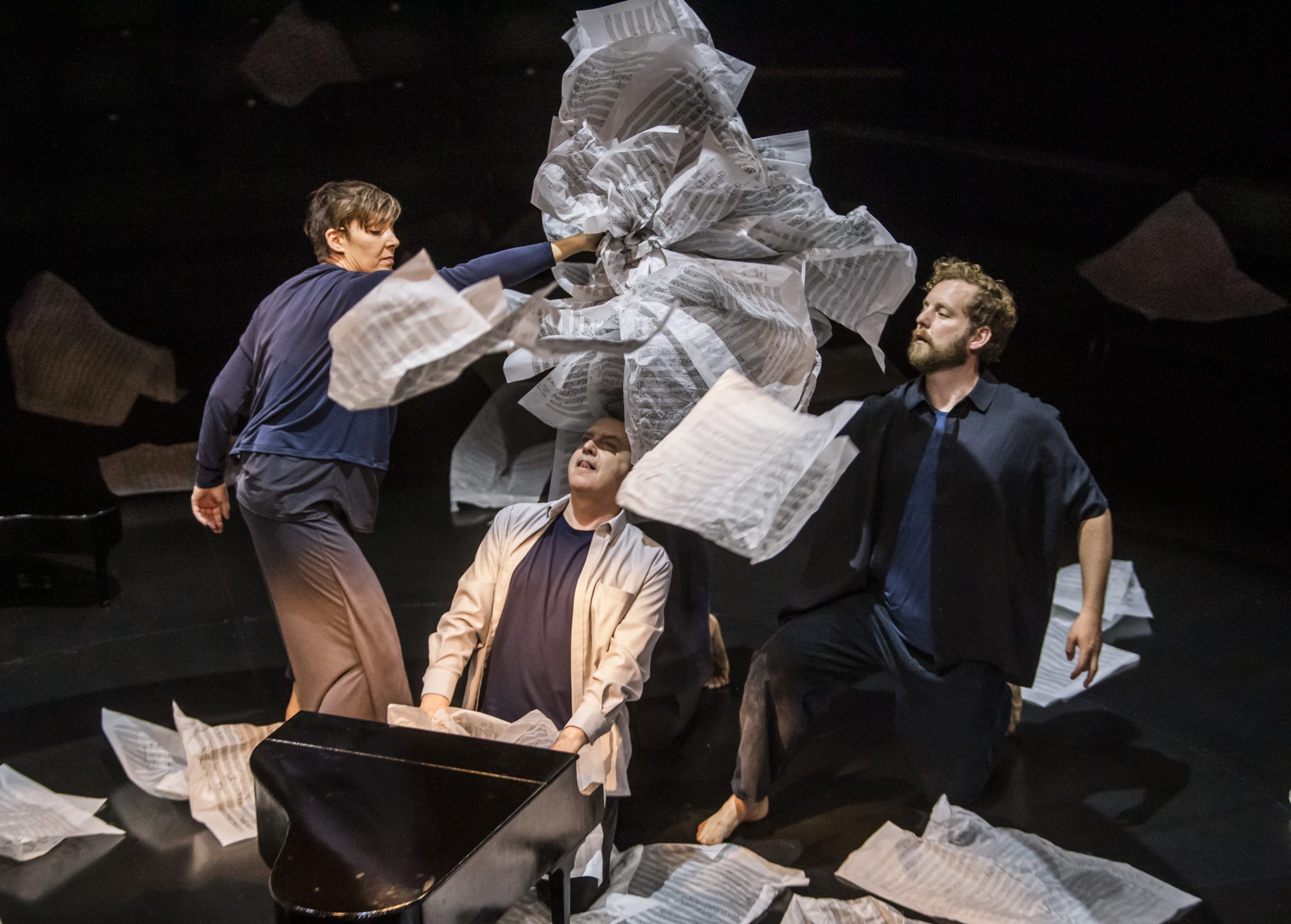Three performers surrounded by scattered sheets of music, one tossing a bundle of paper dramatically in the air. A black piano is partly visible. The scene is dynamic and expressive, set against a dark background.