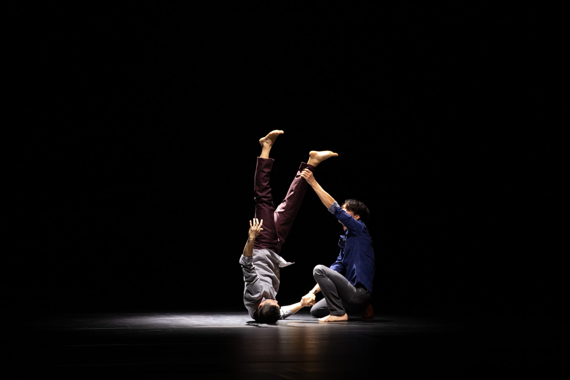 Two dancers perform on a dark stage; one lies on the floor with legs raised vertically while the other kneels, holding their legs. A spotlight highlights them against the black background.