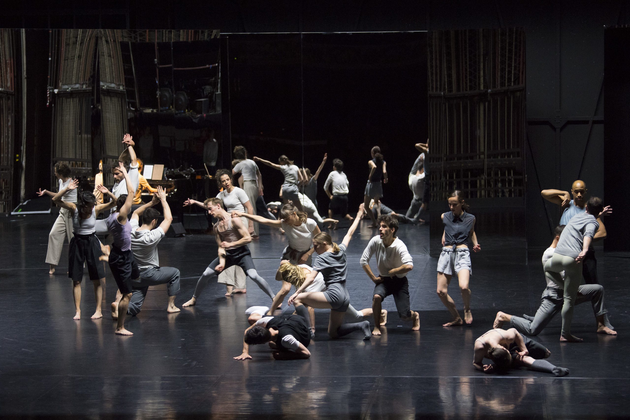 A group of dancers perform an expressive, modern dance routine on a dimly lit stage, with some standing, some crouching, and others mid-motion. The background includes mirrors and stage equipment.