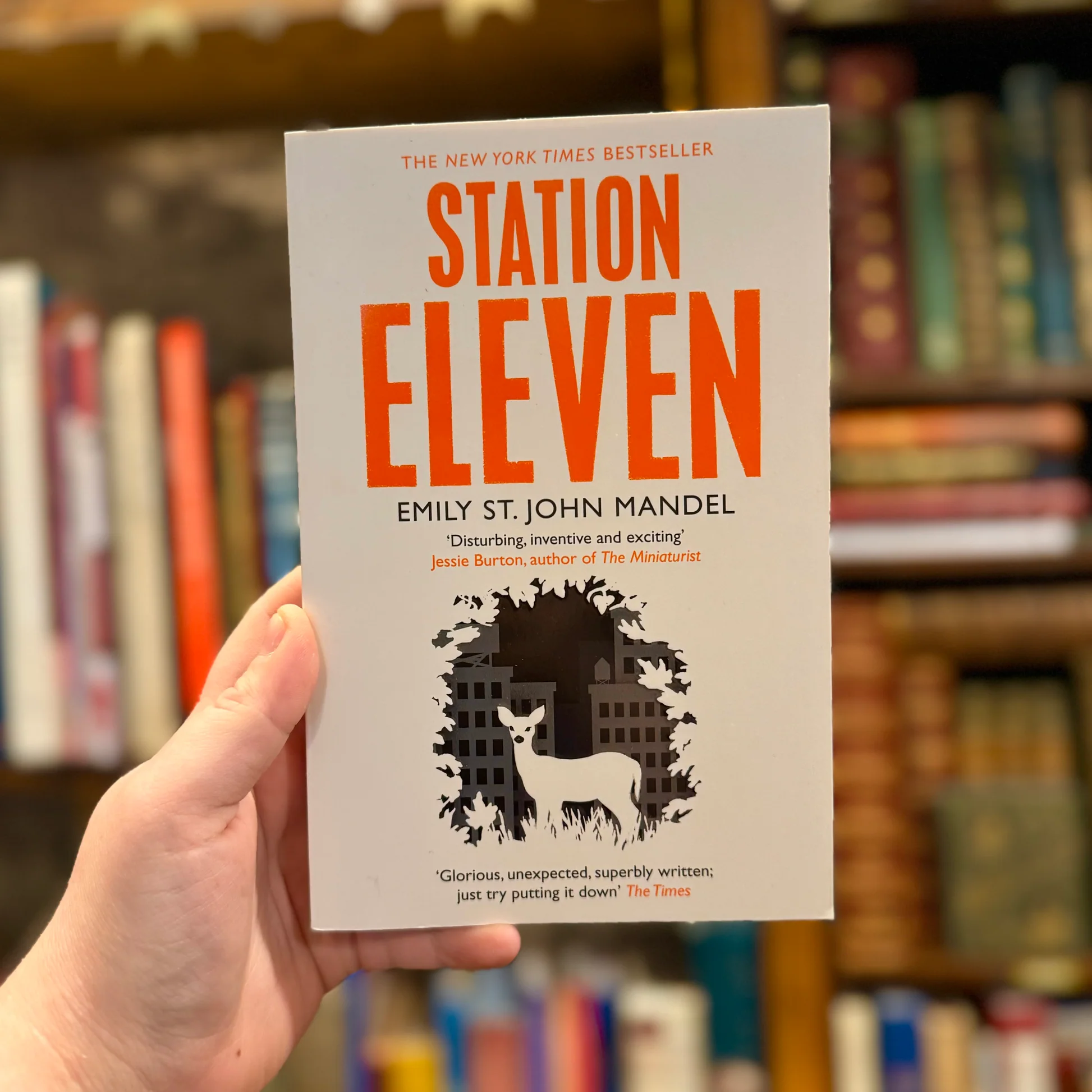 A hand holds the novel “Station Eleven” by Emily St. John Mandel in front of bookshelves. The cover features an orange title and a white deer standing among black silhouetted trees and city buildings.