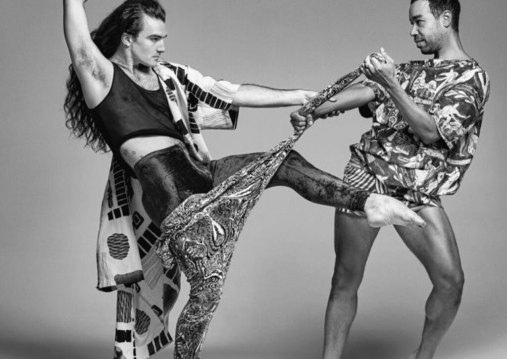 Two people pose dynamically; one raises a leg while the other pulls on patterned fabric. Both wear bold, artistic clothing. The background is plain and the image is in black and white.