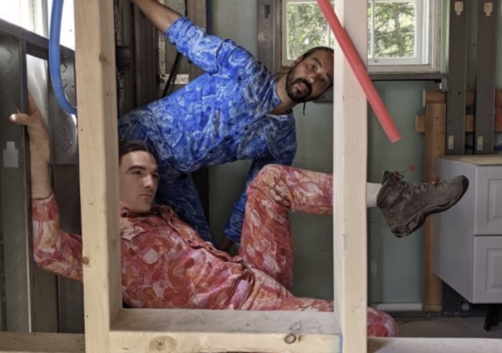 Two people wearing colorful patterned jumpsuits, one blue and one red, pose playfully inside a wooden frame in a room under construction. Light comes in through a window behind them.