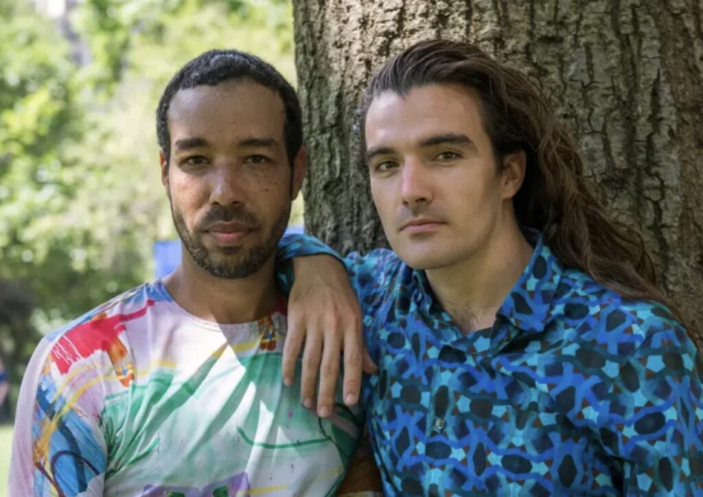 Two people stand close together outdoors by a tree. One rests their arm on the other’s shoulder. Both look at the camera, surrounded by green foliage and sunlight.