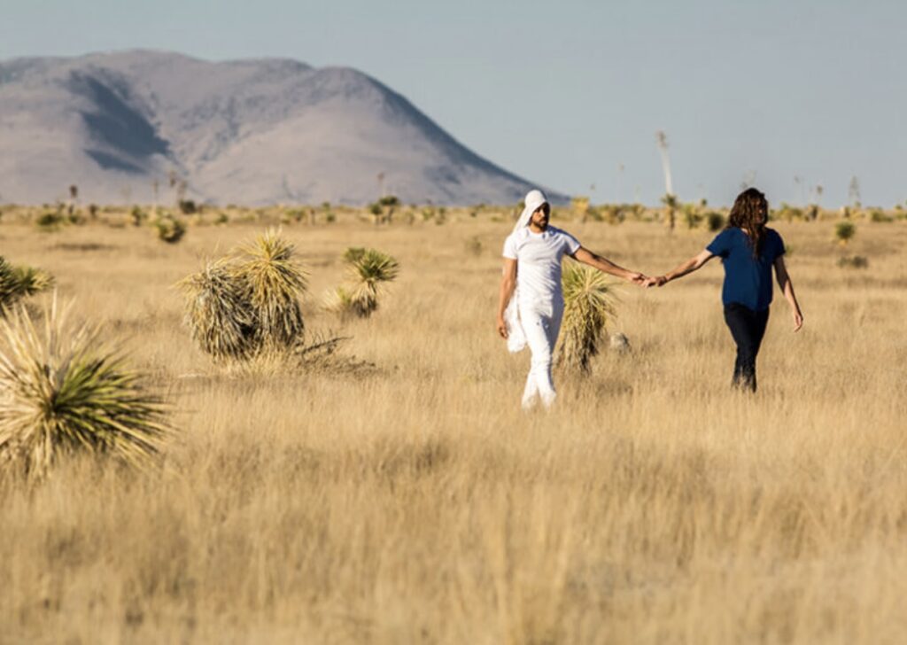 Two people holding hands walk through a dry grassy field with scattered bushes, mountains visible in the background under a clear sky.