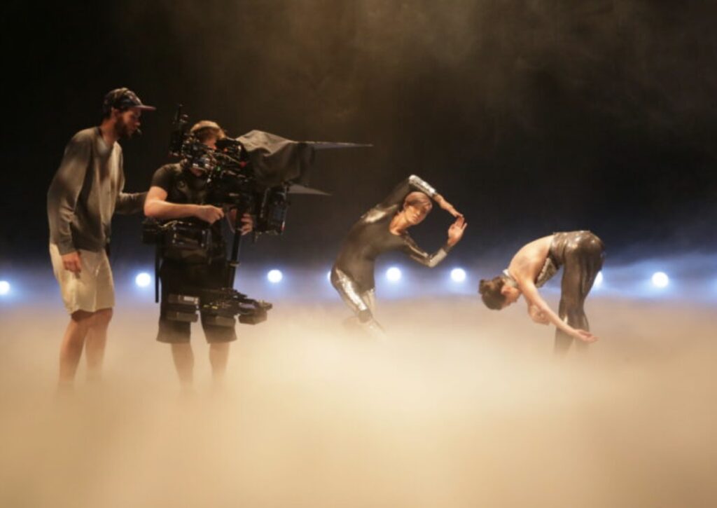 A camera crew films two dancers in metallic costumes performing expressive poses on a misty stage, with soft lights glowing in the background.
