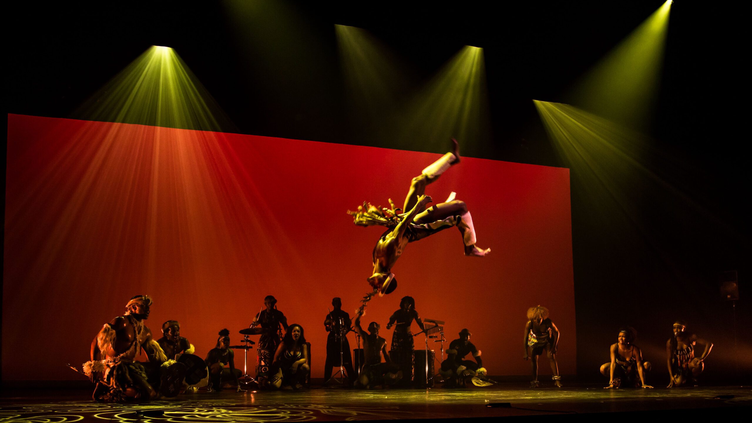 An acrobat performs a mid-air flip onstage, surrounded by musicians and dancers under dramatic yellow spotlights, with a vibrant orange-red backdrop behind them.