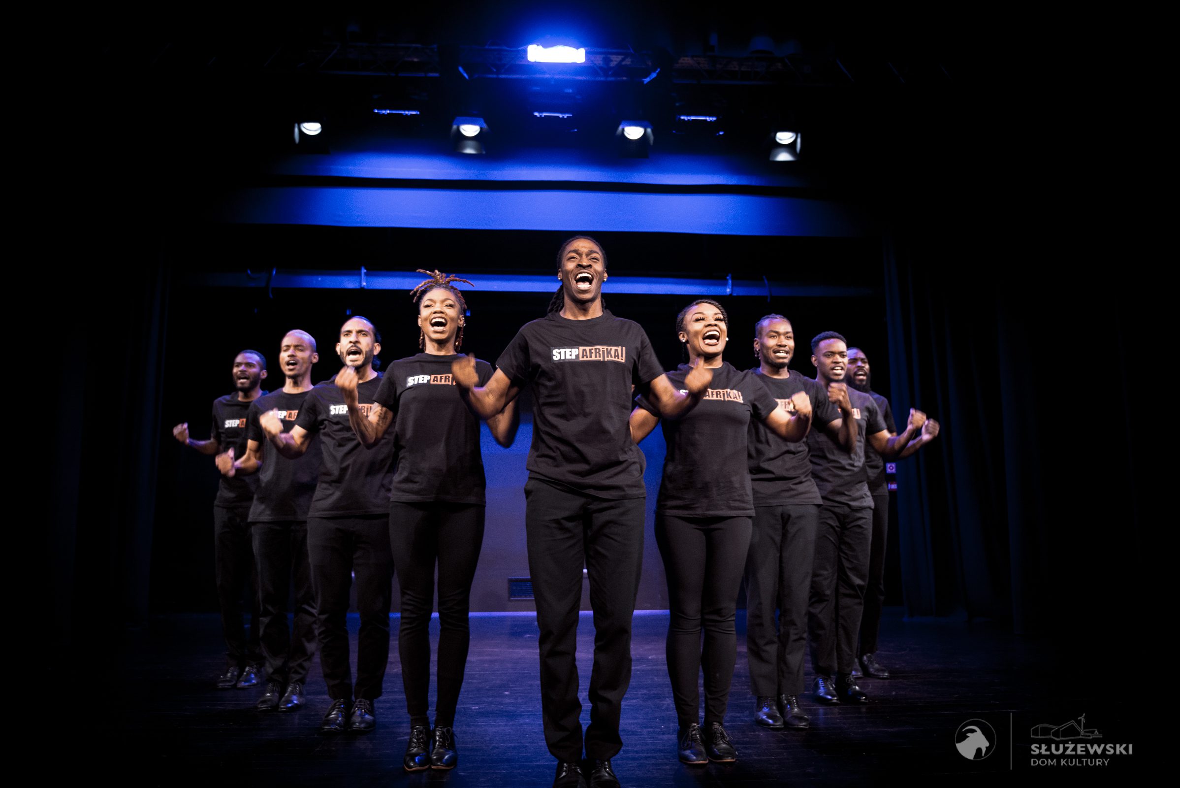 A group of performers in black “STEP AFRIKA!” shirts smile and clap in unison on stage, with bright stage lights and a blue-lit background behind them.