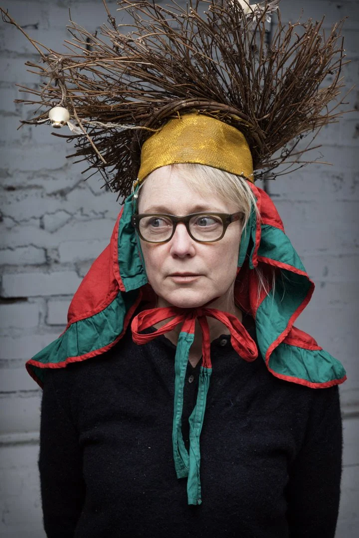 A woman wearing glasses, a gold headpiece with twigs and small lights, and a red and green hooded cape stands against a white brick wall, looking to the side.