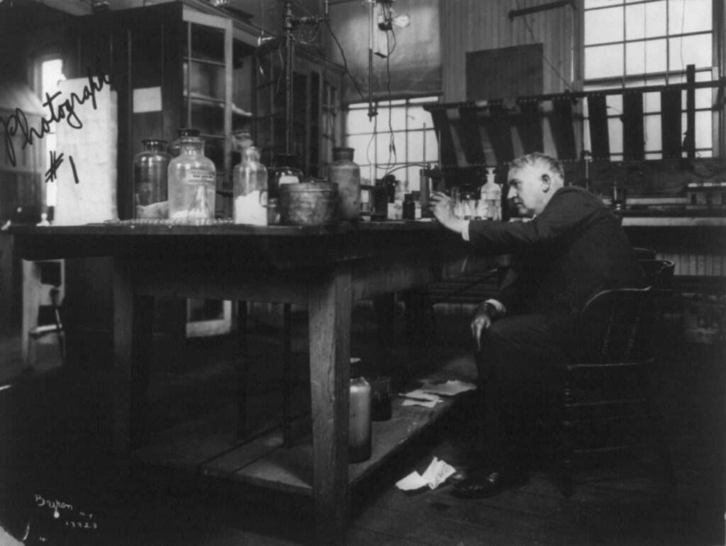 An older man sits at a cluttered laboratory table examining a glass flask, surrounded by bottles and scientific equipment, in a room with large windows and wooden furniture.