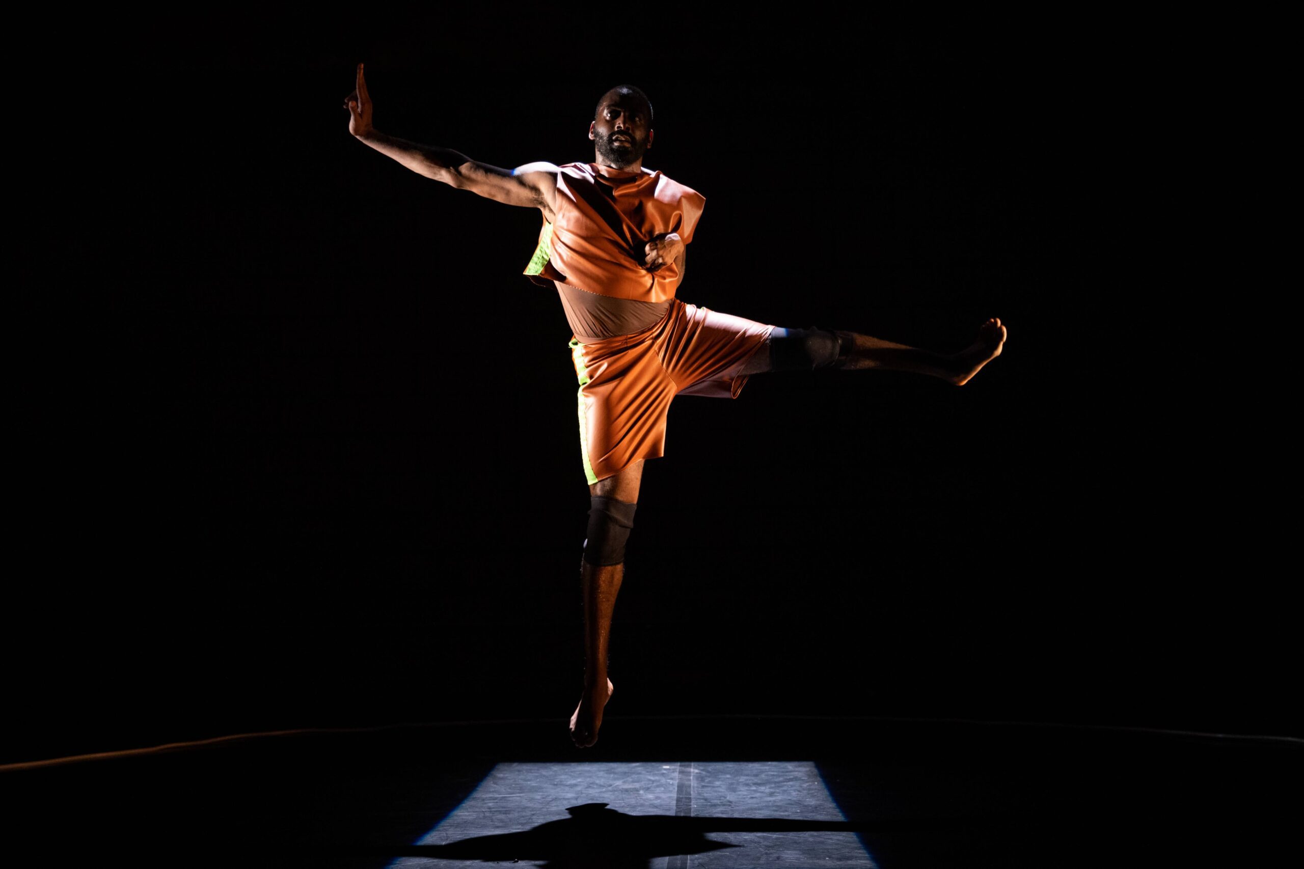 A dancer in orange costume leaps mid-air on a dark stage, spotlight illuminating him, with one arm and one leg extended outward in a dynamic pose.