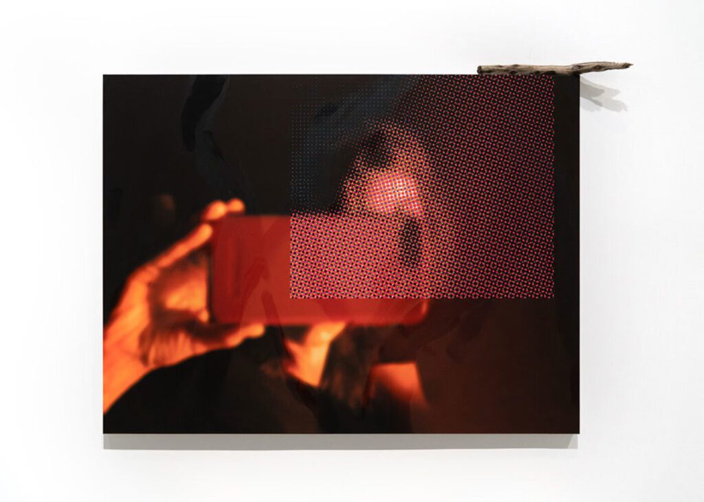 A blurry reflection of a person taking a photo with a phone appears on a dark, glossy surface. Red dots form a rectangular pattern beside their face. A piece of cardboard sits on the upper edge.