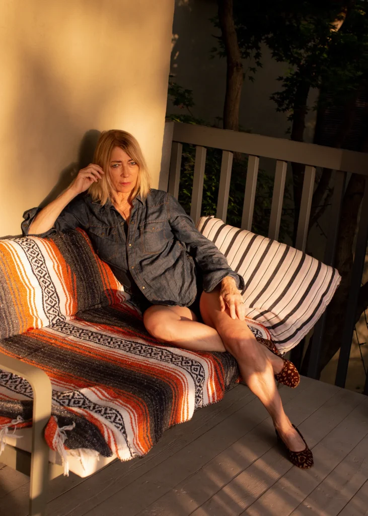 A woman with blonde hair sits on a striped blanket-covered outdoor bench, wearing a denim shirt and patterned flats. Sunlight casts warm shadows on the porch and her face.