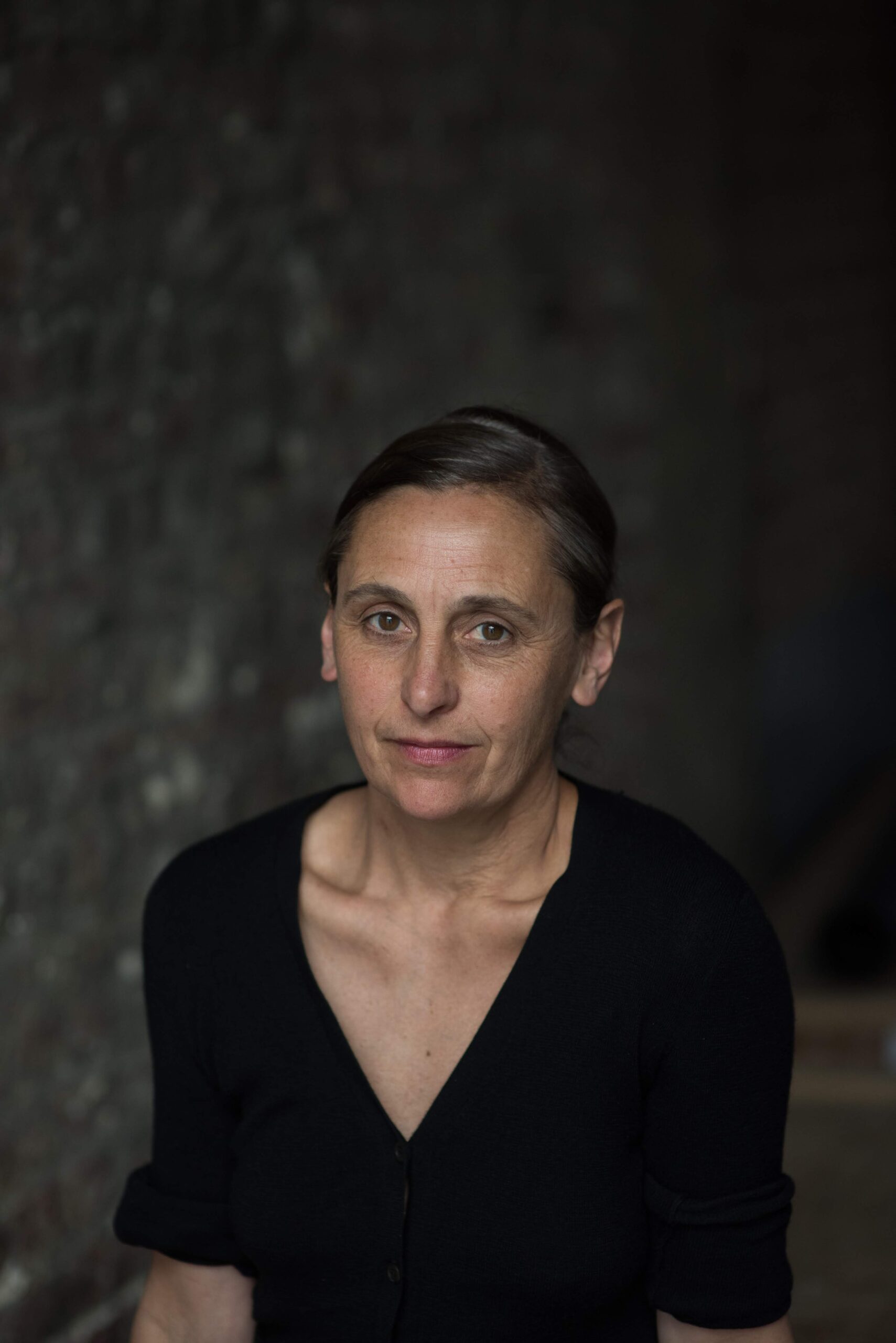 A woman with dark hair pulled back wears a black top. She gazes softly at the camera, standing against a blurred, textured wall with a calm, neutral expression.