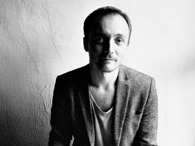 Black and white photo of a man wearing a blazer over a T-shirt, sitting against a textured wall, looking into the camera with a slight smile.