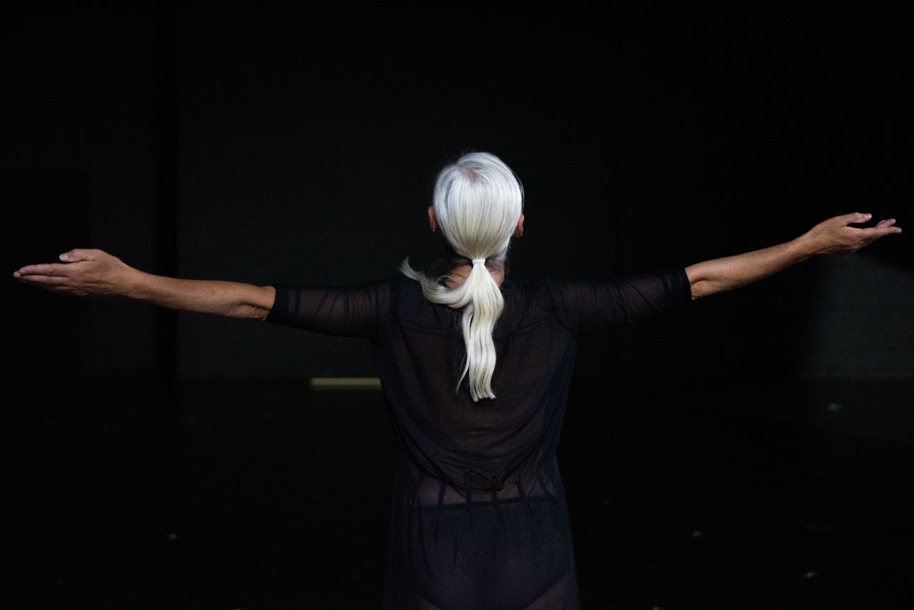 A person with long white hair stands with arms outstretched in a dark space, viewed from behind, wearing a sheer black top.