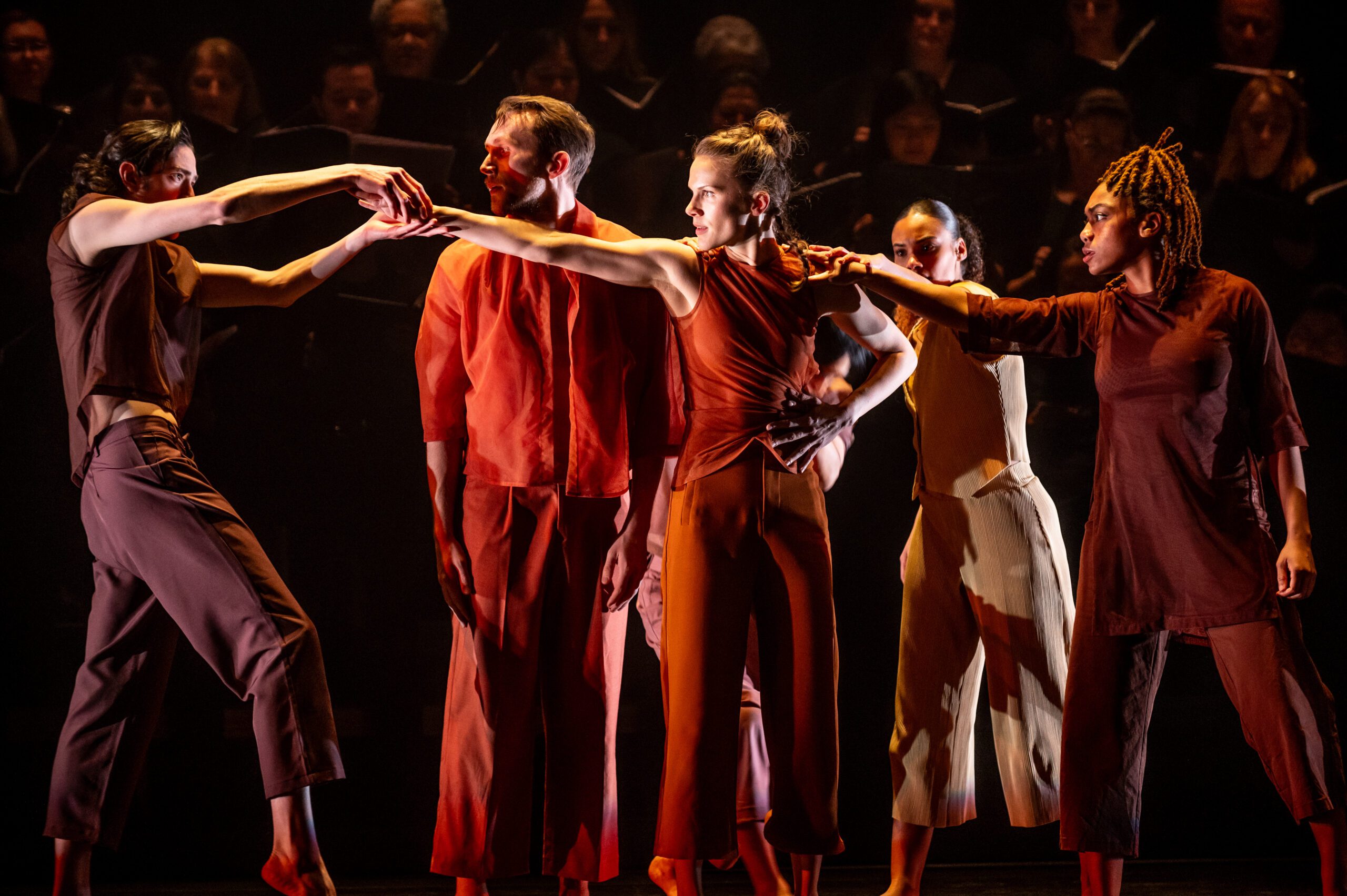 Five dancers in earth-toned outfits stand closely together on stage, their arms interlinked in an expressive pose, with a choir visible in the dimly lit background.