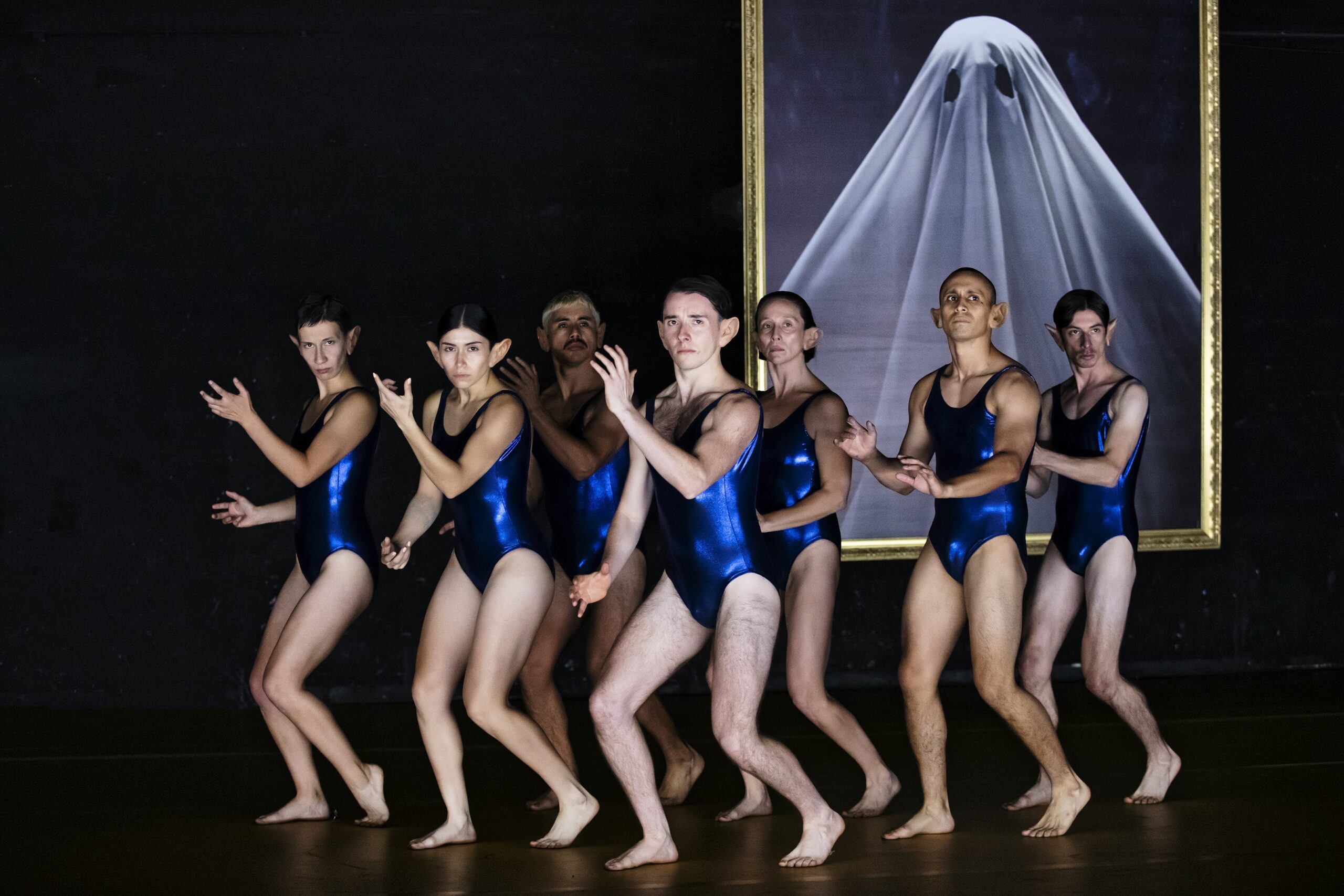 Six people in blue leotards pose dramatically in front of a framed photo of a ghostly white sheet figure on a dark wall.