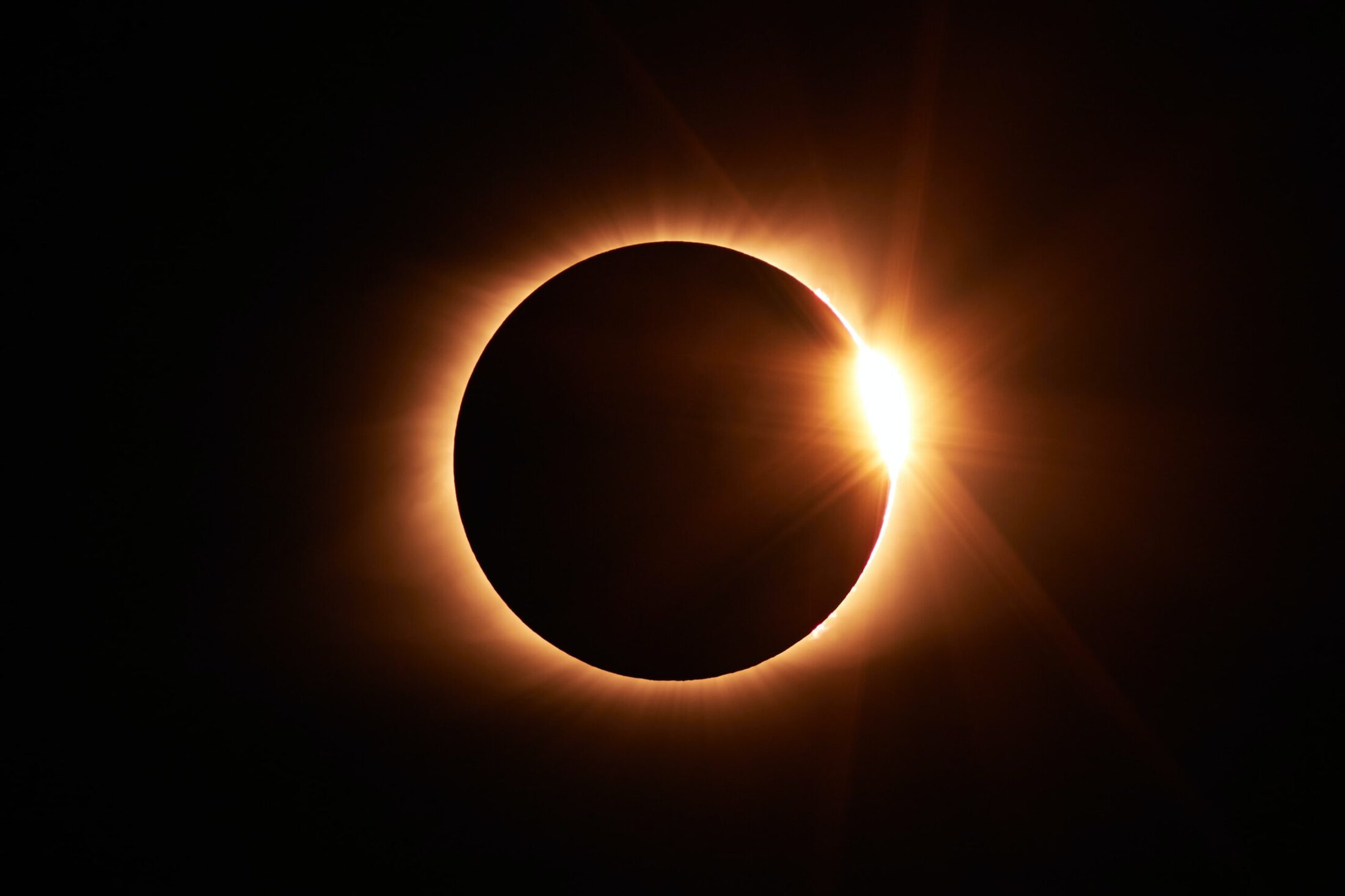 A total solar eclipse with the sun’s corona glowing around the dark moon; a bright burst of light appears at the edge, known as the “diamond ring” effect. The background is pitch black.