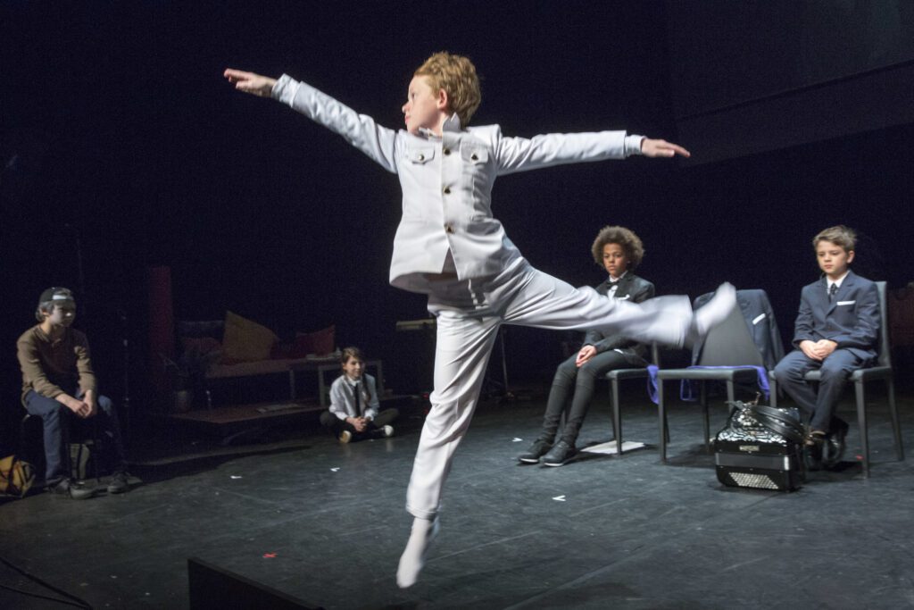 A child in a light suit leaps gracefully across a stage, while four other children, dressed formally, sit and watch in the background.