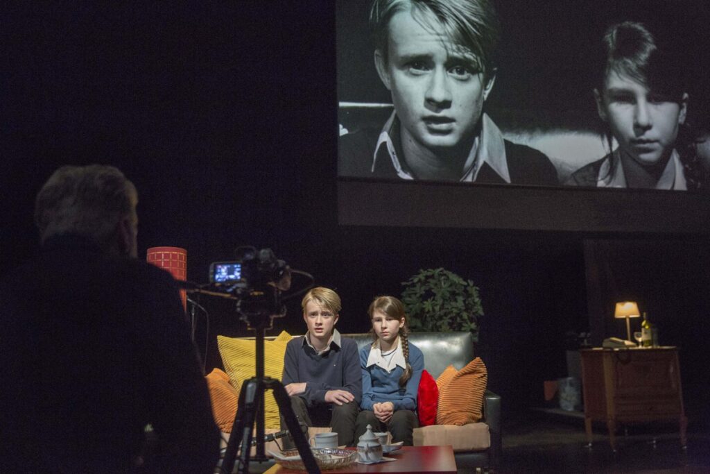 Two children sit on a sofa being filmed, with their black-and-white image projected above them. A man operates the camera, and the room is furnished with lamps, pillows, and a side table.