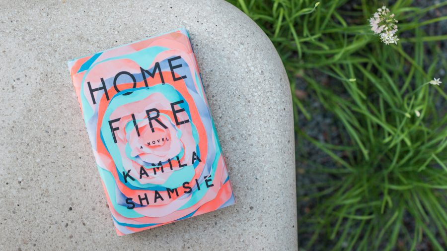 The book “Home Fire” by Kamila Shamsie rests on a curved concrete surface, with green plants and a small white flower visible nearby.