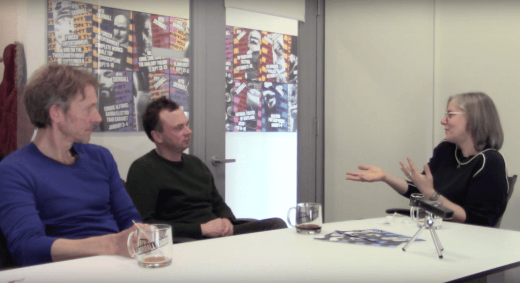 Three people sit around a table having a discussion. Two men are on the left, facing a woman on the right who gestures with her hands. Posters are visible on the wall behind them. Cups and papers are on the table.