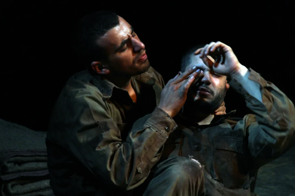 Two men in worn, dirty clothing sit close together. One gently tends to the other’s eye, both appearing serious and focused, suggesting a moment of care or aid in a difficult situation. The background is dark.