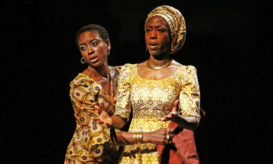 Two women in patterned dresses and gold jewelry stand close together on stage, one comforting the other. The background is dark, highlighting their expressive faces and gestures.