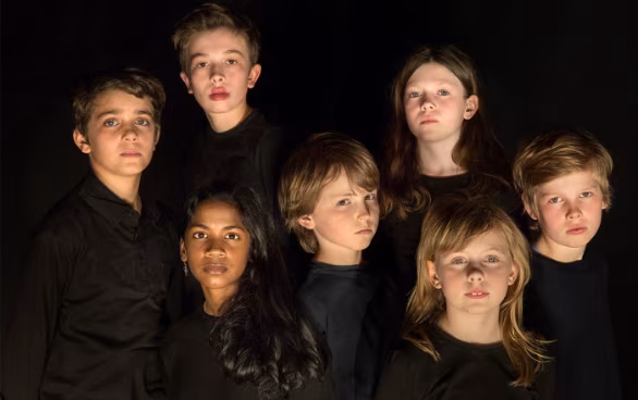 Seven children in black shirts stand together against a dark background, looking directly at the camera with serious expressions.
