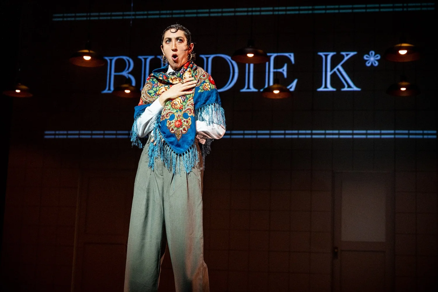 A person wearing a colorful shawl and loose pants stands on stage, hand on chest, with a surprised expression. The background shows the lit-up partial text “BLUDIE K*” on a tiled wall.