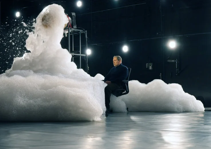 A man sits in a chair surrounded by a large cloud of white foam, with bright lights illuminating the scene in a dark room.