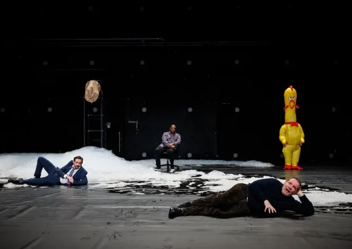 Three men are on a dark stage; two lie on the floor, one is seated. Snow-like piles are onstage. An inflatable yellow chicken stands at the back. The setting is surreal and theatrical.