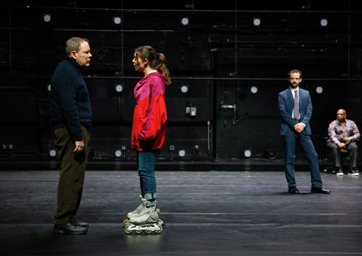A woman in a red jacket and rollerblades faces a man in a dark sweater on a stage, while two men in formal clothes watch from the background.