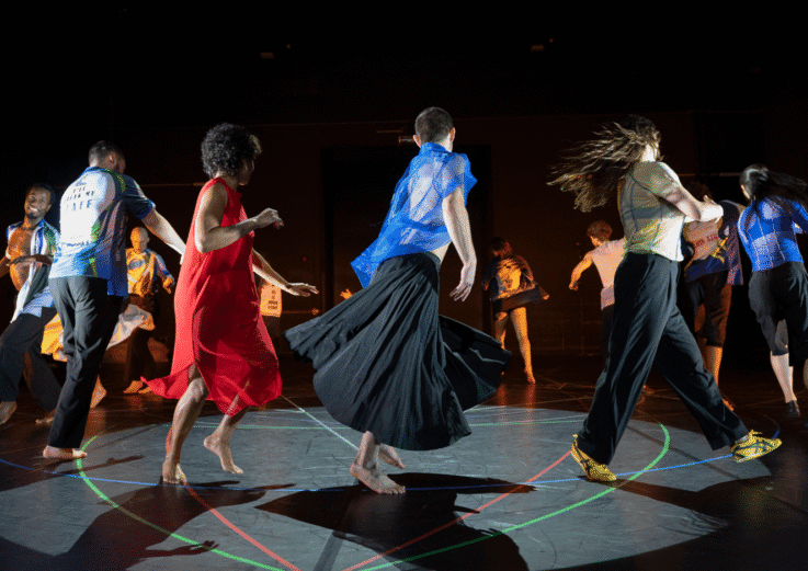 A group of dancers in colorful costumes perform barefoot on a dark stage, moving in a circle with energetic poses under bright lighting and neon lines on the floor.