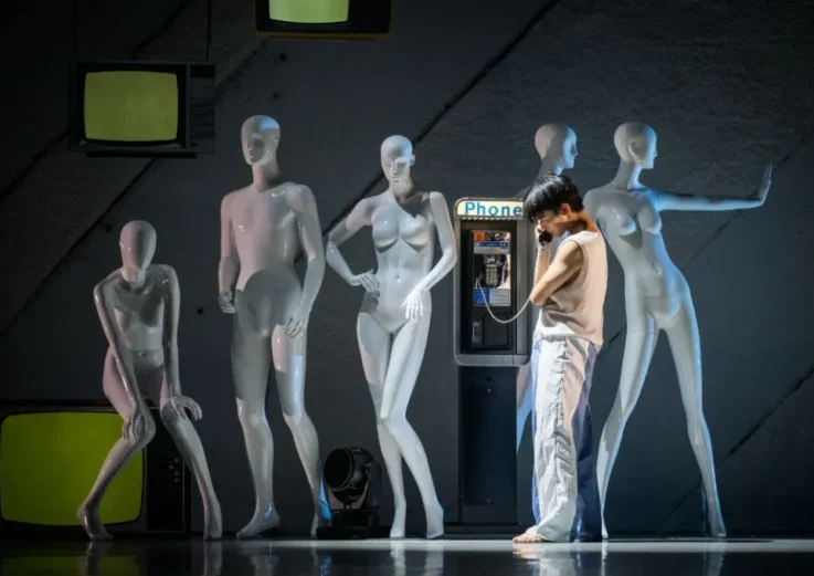 A person stands barefoot at a payphone, surrounded by five white mannequins in various poses; old TVs are in the background, creating a surreal scene.