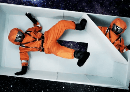 Two people in orange space suits lie in separate white, box-like compartments against a starry outer space background.