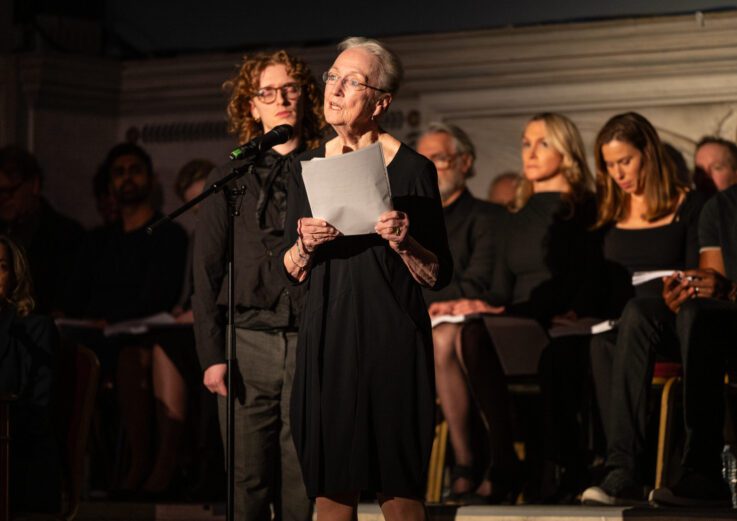 An older woman reads from a paper at a microphone on stage, with people seated behind her, all dressed in black.
