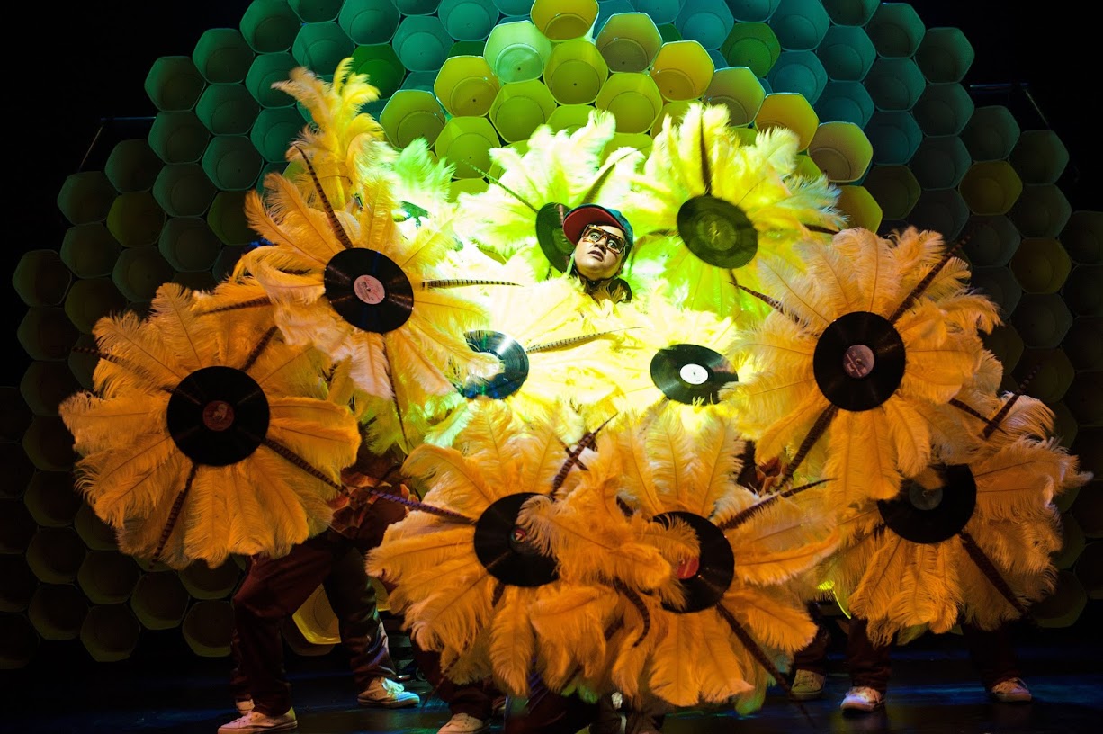 A performer stands in front of a honeycomb-like backdrop, surrounded by people holding large yellow feather fans with vinyl records at their centers, creating a vibrant and dynamic stage scene.