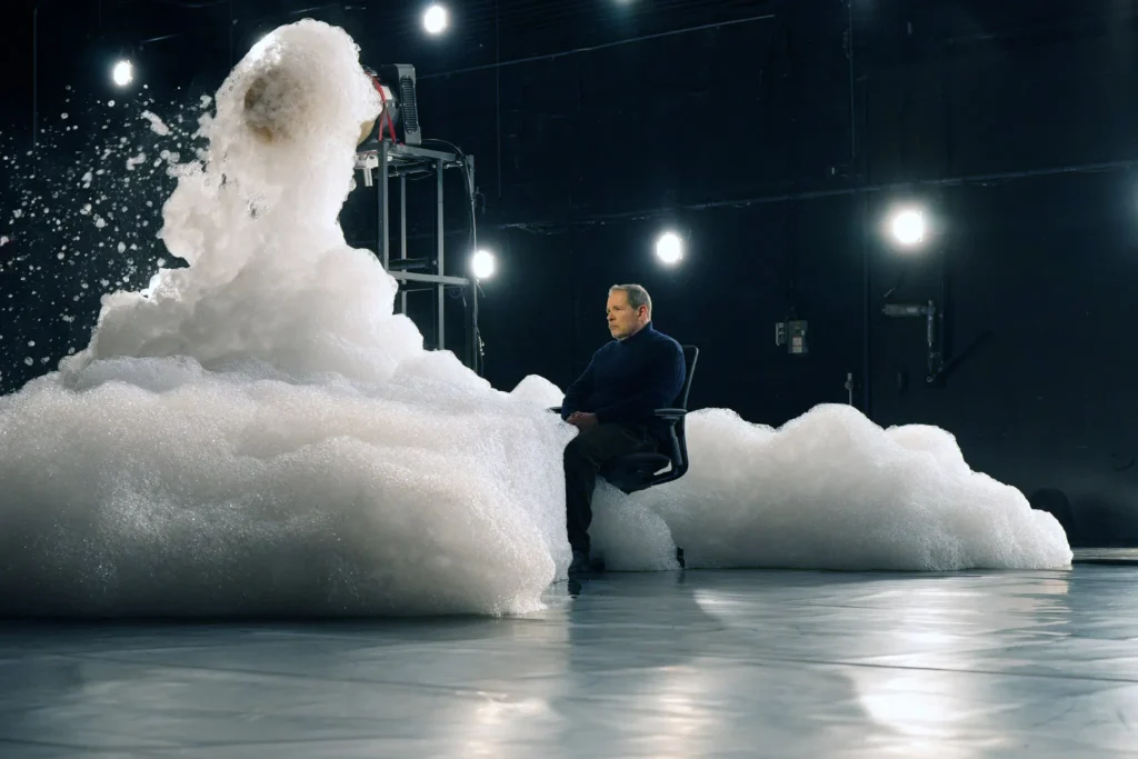 A person in a chair sits calmly as a large burst of foam and bubbles erupts beside them in a dimly lit room with bright lights on the walls.