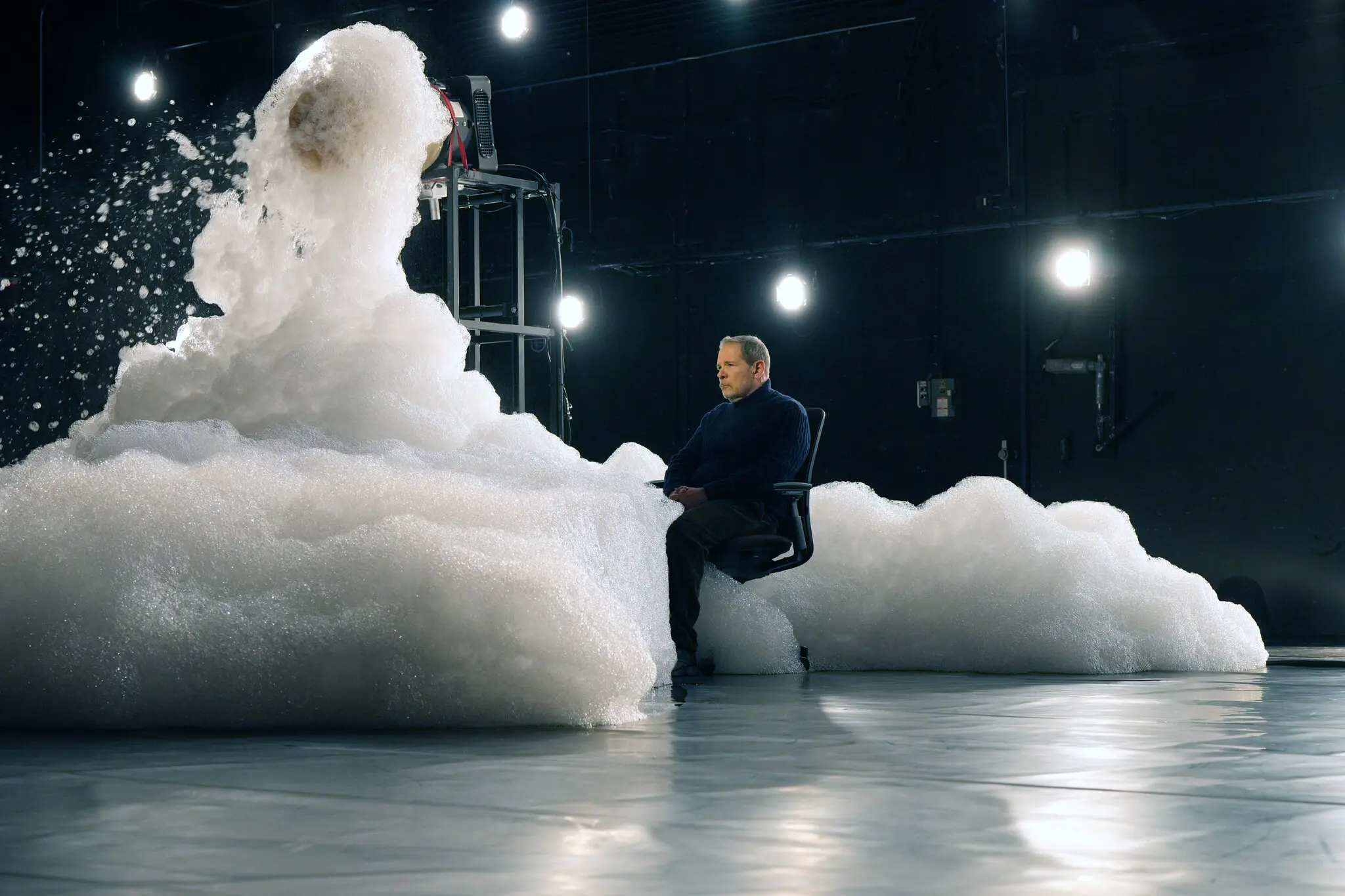 A person in a chair sits calmly as a large burst of foam and bubbles erupts beside them in a dimly lit room with bright lights on the walls.