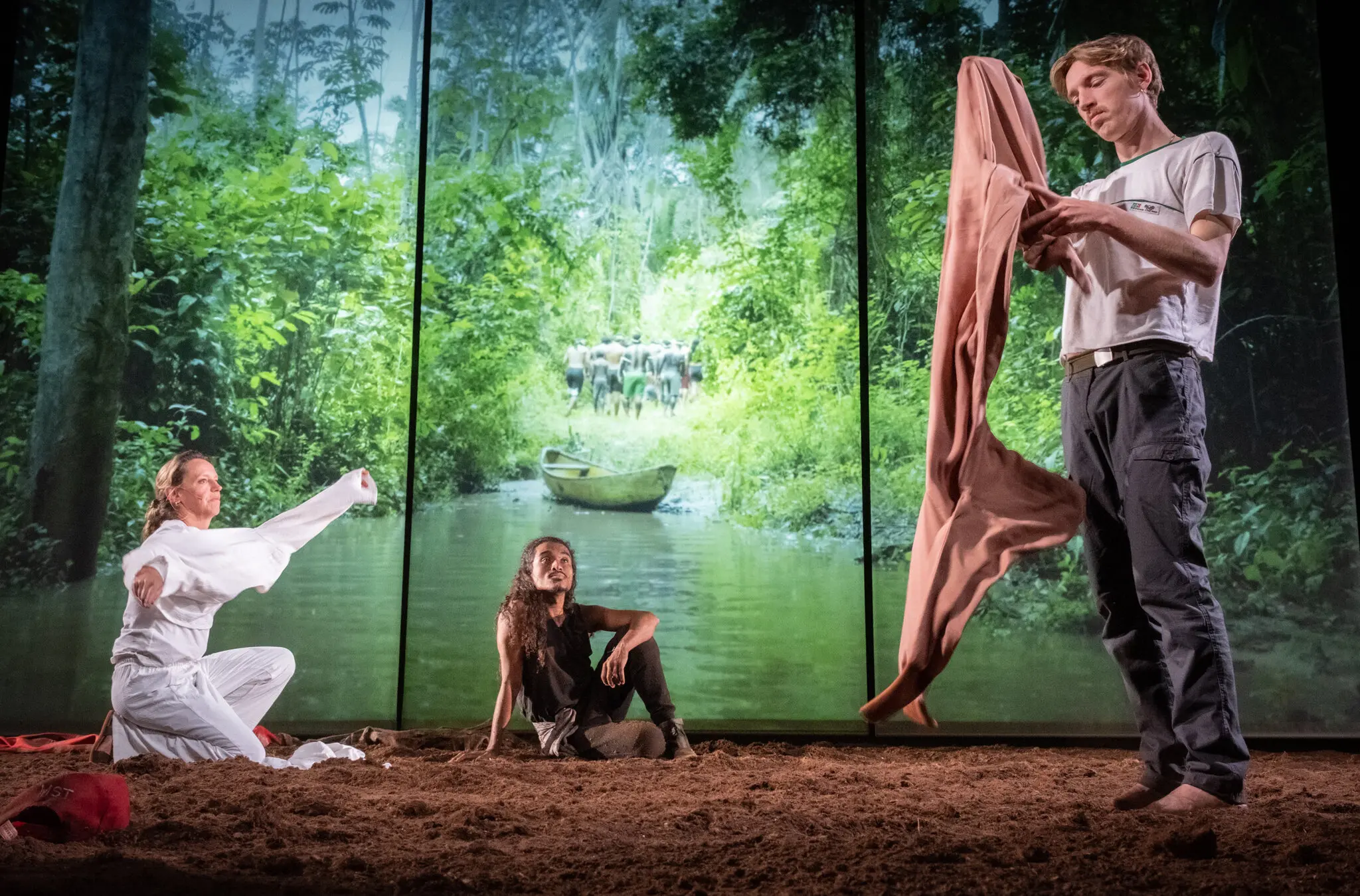 Three people perform onstage with a forest and river projected behind them. One kneels with arms raised, another sits smiling, and a third stands holding clothing. The ground is brown, resembling dirt.