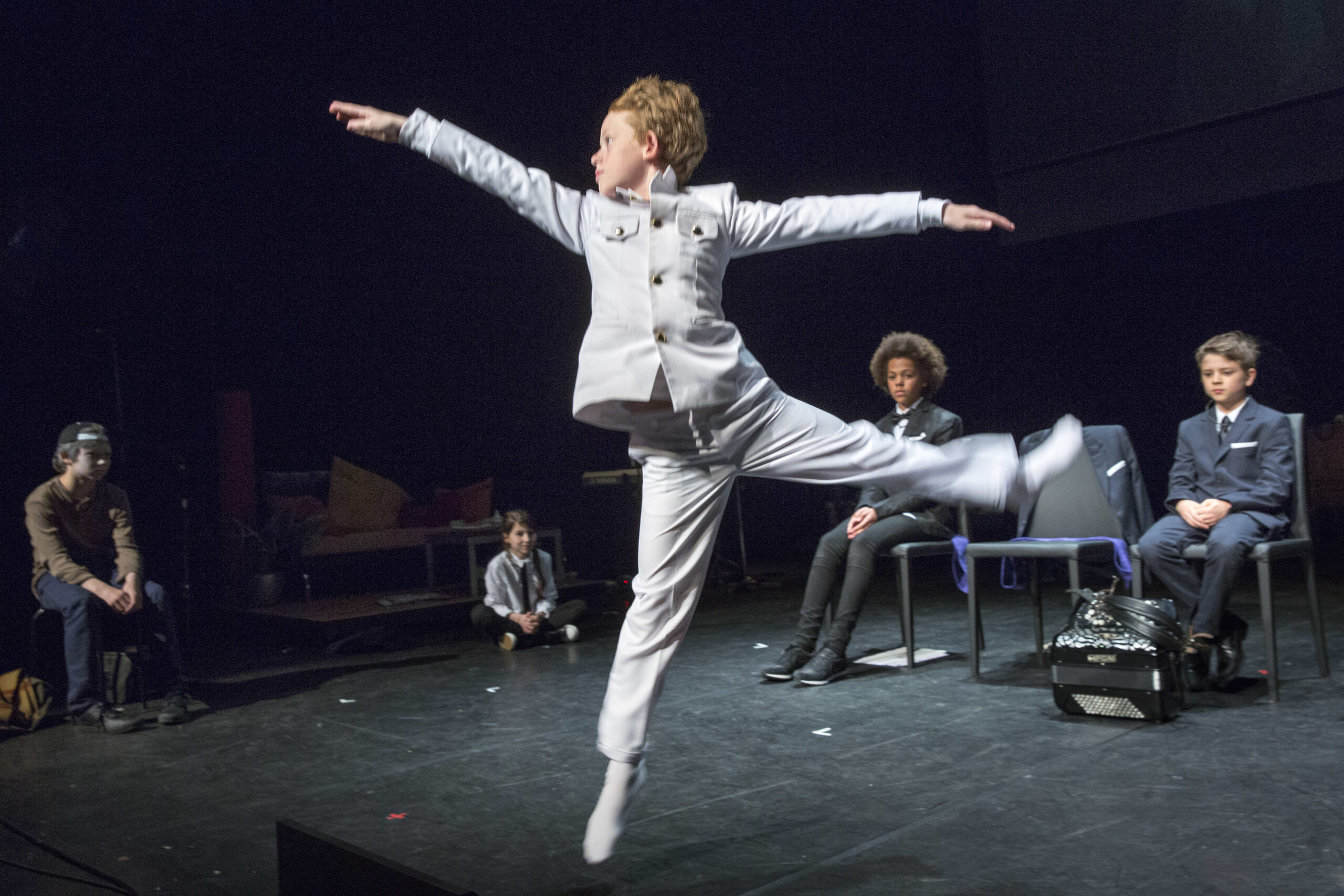 A child in a light suit leaps mid-air on stage, arms outstretched, while four other children in darker suits watch, seated in the background.