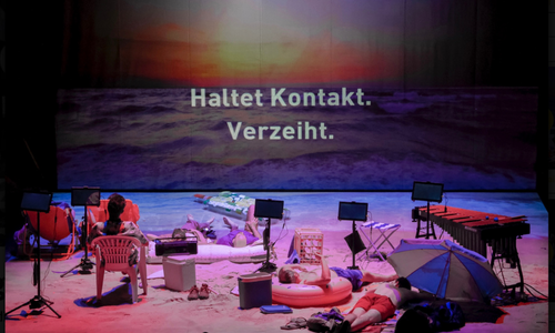 A stage set like a beach shows people resting among props. A large screen behind them displays a sunset over the ocean and German text: “Haltet Kontakt. Verzeiht.” (Stay in touch. Forgive.).
