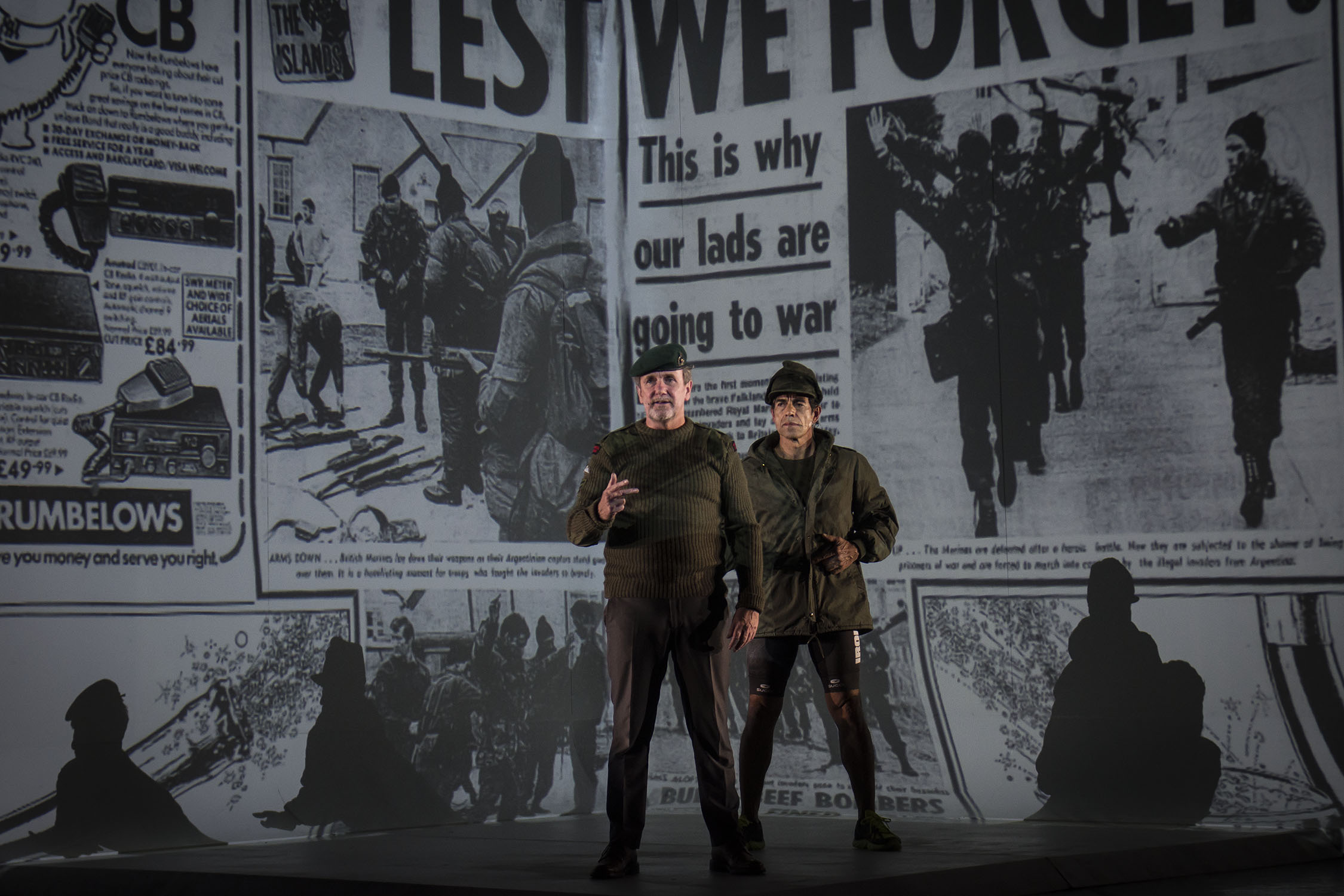 Two actors dressed as soldiers stand on stage, with a large black-and-white newspaper collage about war projected behind them. The headlines read, “LEST WE FORGET” and “This is why our lads are going to war.”.