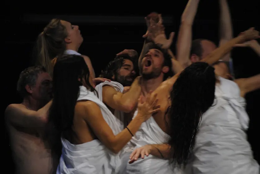 A group of people wrapped in white sheets are reaching and interacting with expressive gestures against a dark background, suggesting a dramatic or theatrical scene.