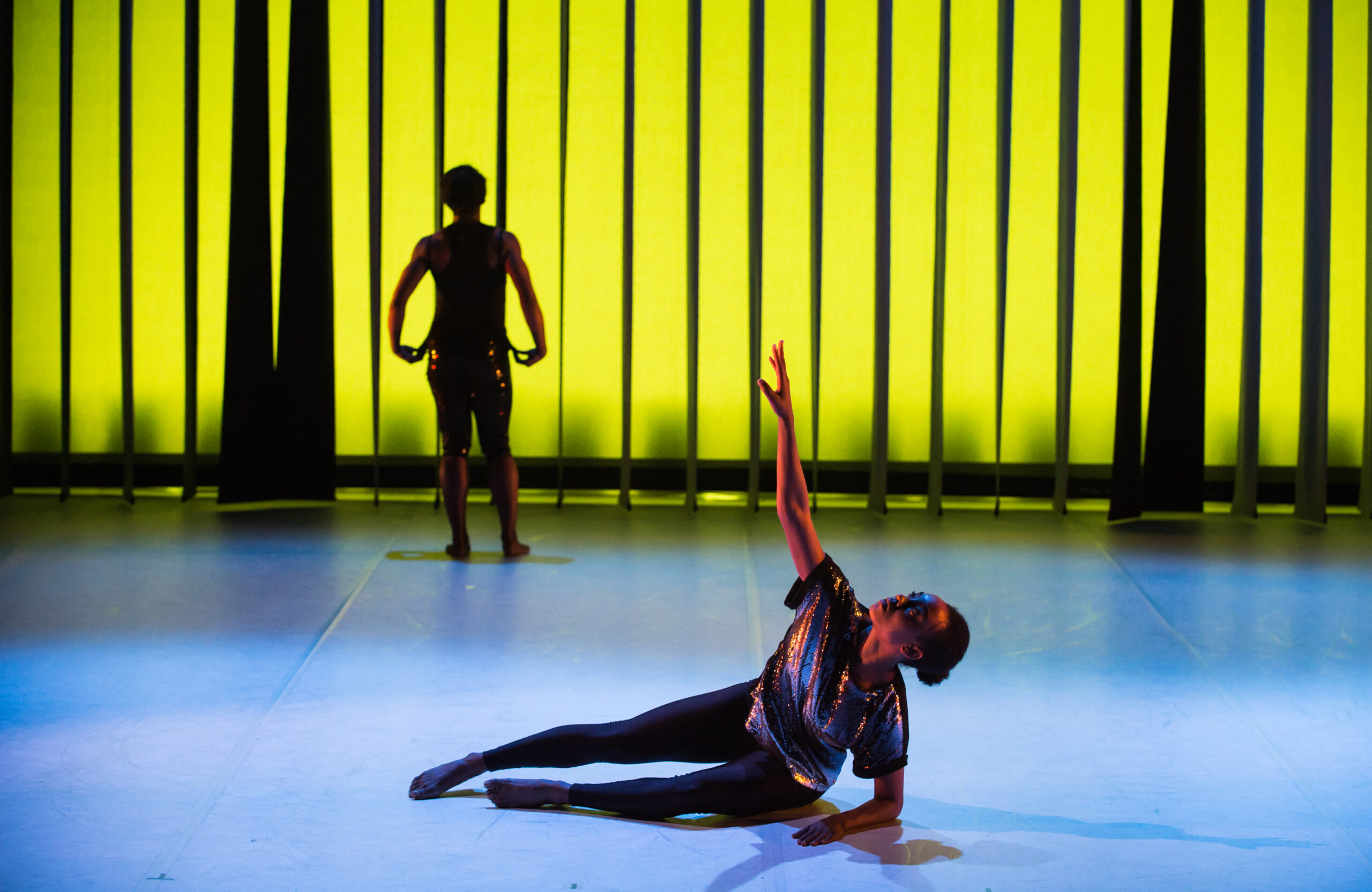 A dancer in black and silver lies on the floor reaching upward, while another dancer stands facing a glowing yellow backdrop with vertical black bars.