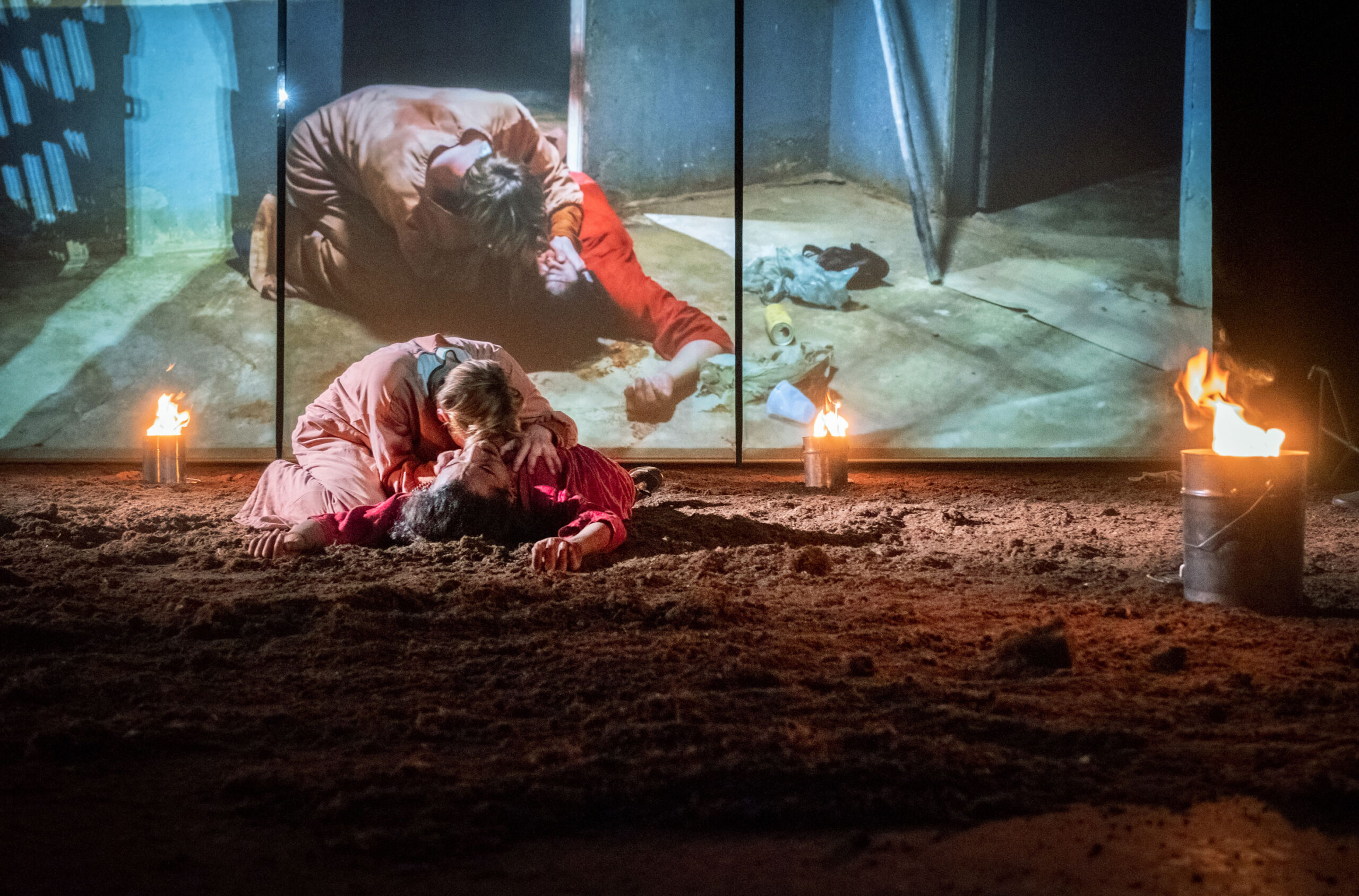 Two people in dramatic costumes lie on a sandy stage with fire torches and a metal bucket. A projection behind them shows a similar scene, adding intensity to the performance.