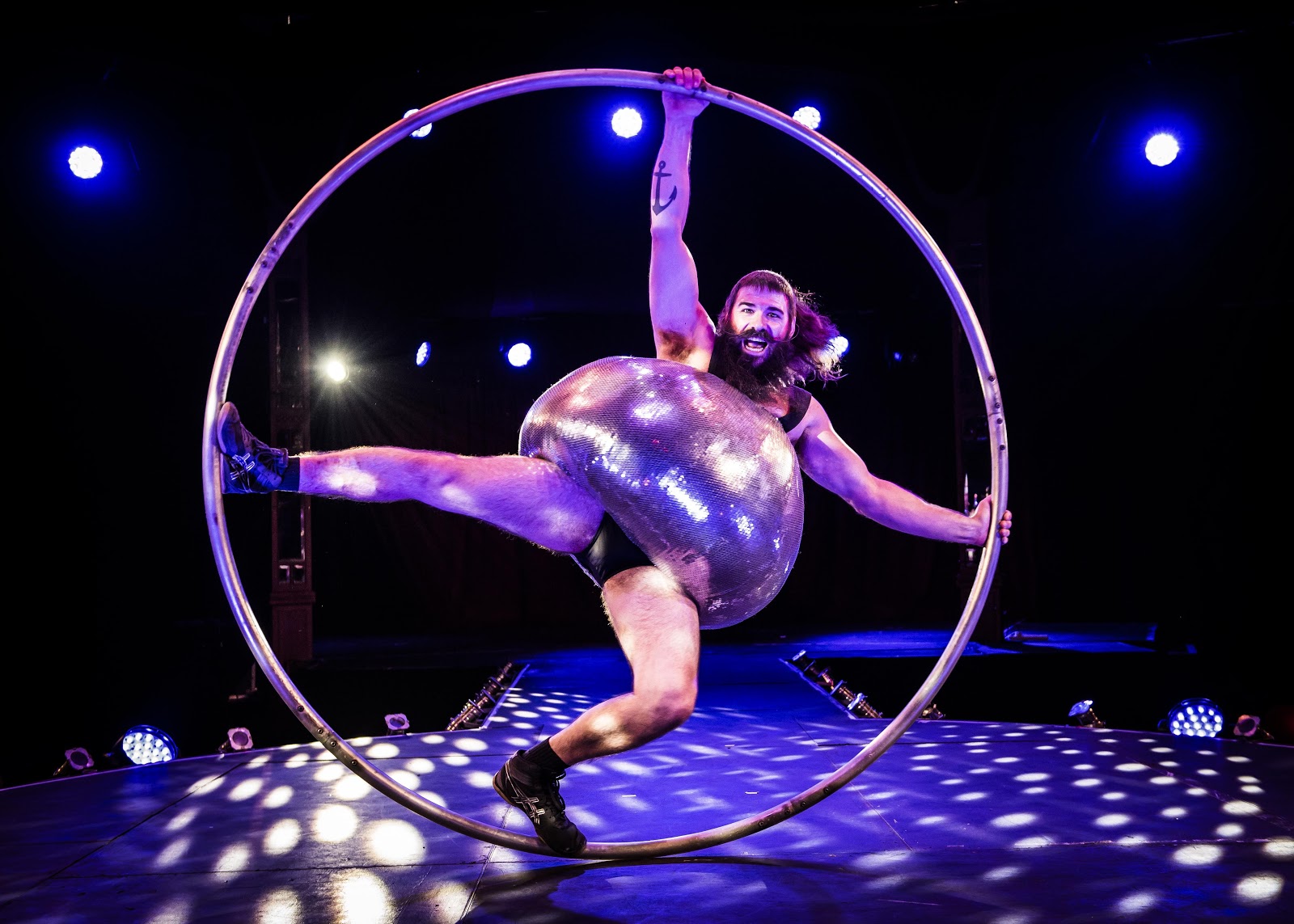 A performer with a sparkling, oversized, round costume poses inside a large metal hoop on stage, lit by blue and purple spotlights.
